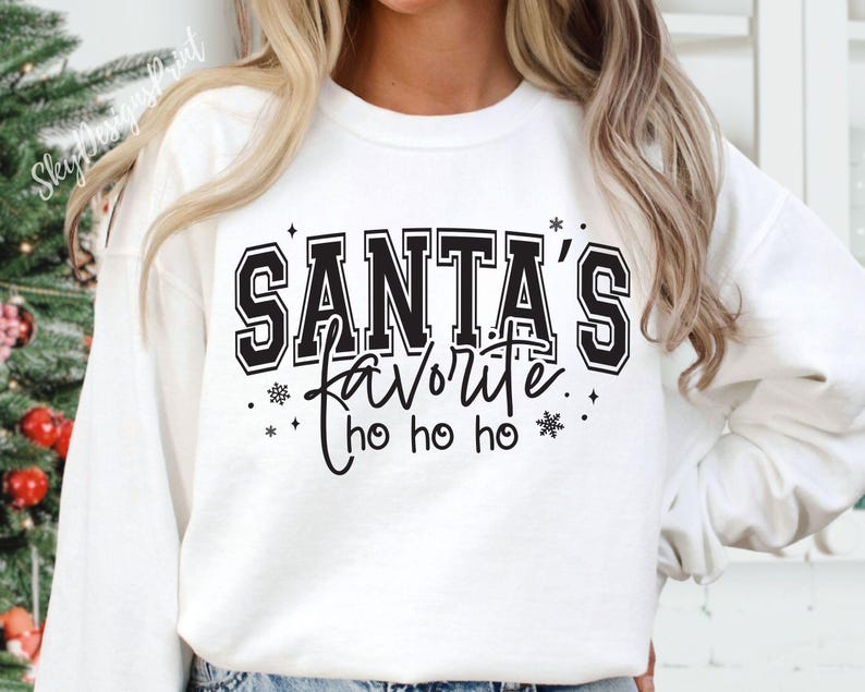 May include: White sweatshirt with the text "SANTA'S FAVORITE" in black block letters, with "ho ho ho" below. The design includes snowflakes and stars. A festive holiday-themed top.