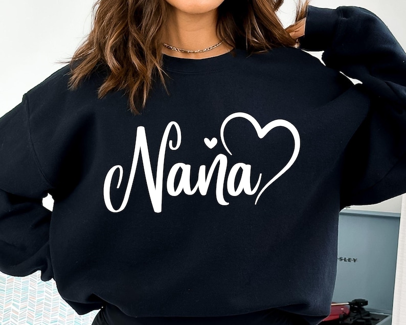 Puede incluir: Sudadera negra con la palabra "Nana" en letras blancas cursivas y un dise&ntilde;o de coraz&oacute;n blanco.