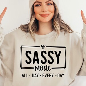 May include: A beige sweatshirt with a black graphic that says "Sassy Mode" with a heart above the word "Sassy" and the words "All Day Every Day" below.