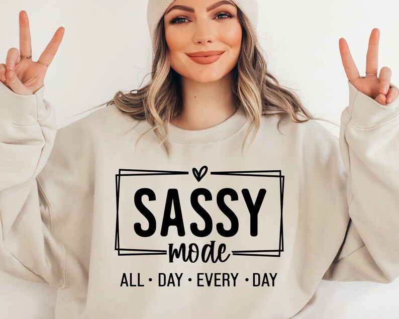 May include: A beige sweatshirt with a black graphic that says "Sassy Mode" with a heart above the word "Sassy" and the words "All Day Every Day" below.