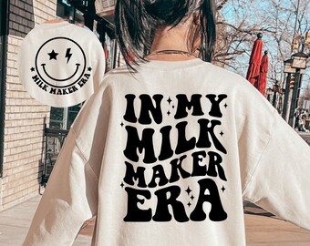 May include: A white sweatshirt with the text "IN MY MILK MAKER ERA" printed in black retro-style lettering. The text is surrounded by small stars and sparkles.