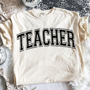 May include: Cream-colored t-shirt with the word "TEACHER" in bold, black, collegiate-style lettering. The shirt is made of a soft material and has a classic crew neck. The design is centered on the front of the shirt.