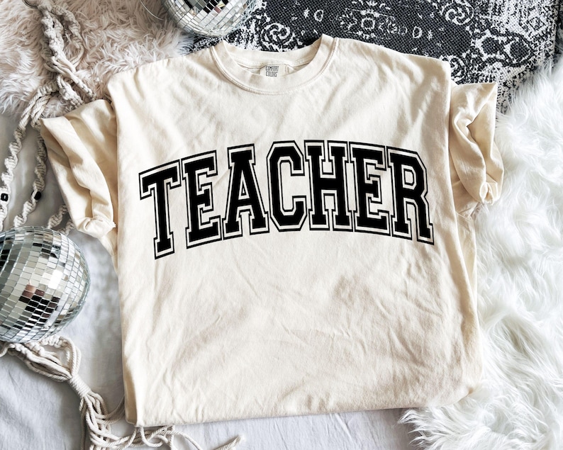May include: Cream-colored t-shirt with the word "TEACHER" in bold, black, collegiate-style lettering. The shirt is made of a soft material and has a classic crew neck. The design is centered on the front of the shirt.