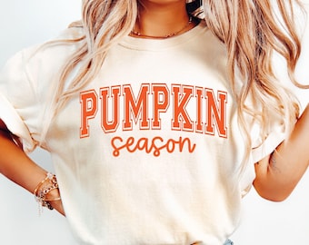 Może przedstawiać: Kremowa koszulka z napisem "PUMPKIN season" w pomarańczowych literach. Słowo "PUMPKIN" jest pogrubione i obramowane, a "season" jest napisane kursywą. Koszulka ma swobodny styl.