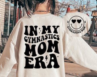 May include: A white sweatshirt with black text that reads "In My Gymnastics Mom Era" and a black smiley face with hearts for eyes that reads "Gymnastics Mom".