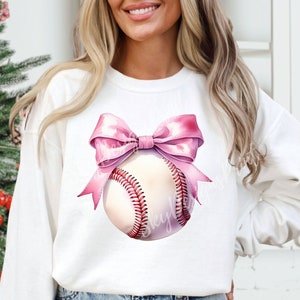 May include: A white sweatshirt with a pink bow and a baseball with red stitching. The baseball is centered on the sweatshirt and the bow is tied around it.