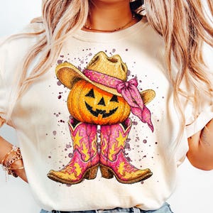 Könnte beinhalten: Ein cremefarbenes T-Shirt mit einer Grafik eines orangefarbenen Kürbisses, der einen beigen Cowboyhut mit einem rosa Bandana trägt. Unter dem Kürbis befinden sich zwei leuchtend rosa Cowboystiefel mit gelben Sternakzenten, umgeben von violetten Farbspritzern.