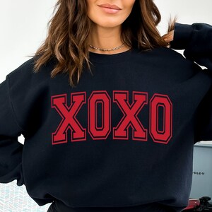 May include: Black sweatshirt with the letters XOXO in red block letters. The letters are outlined in a darker red. The sweatshirt is a crew neck style. The image is a close-up of the sweatshirt.