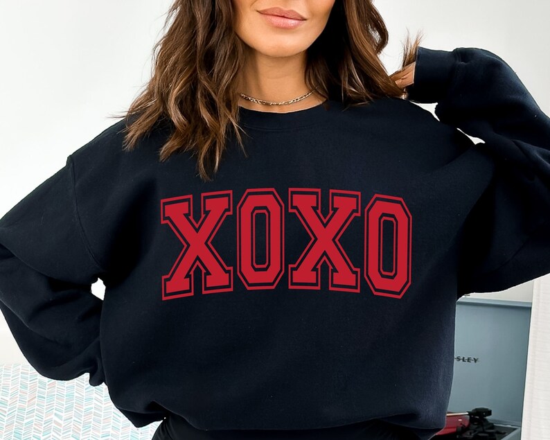 May include: Black sweatshirt with the letters XOXO in red block letters. The letters are outlined in a darker red. The sweatshirt is a crew neck style. The image is a close-up of the sweatshirt.