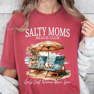 May include: Red t-shirt with the text "Salty Moms Beach Club" and a graphic of two beach chairs under a beach umbrella. The text "Girls Just Wanna Have Sun" is at the bottom of the shirt.