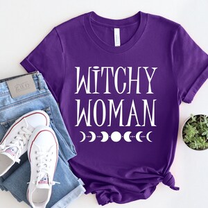 May include: Purple t-shirt with white text that reads "Witchy Woman" and a row of white crescent moons below the text.