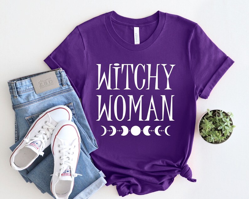 May include: Purple t-shirt with white text that reads "Witchy Woman" and a row of white crescent moons below the text.