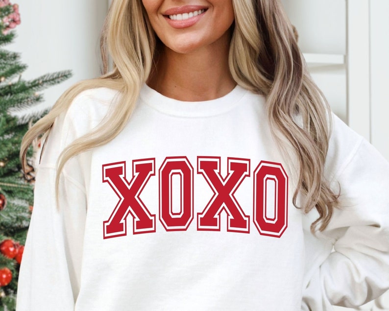May include: White sweatshirt with the word "XOXO" in bold, red, collegiate-style lettering. The letters have a white outline. The sweatshirt is a long-sleeved crew neck. The image is a close-up of the upper body.