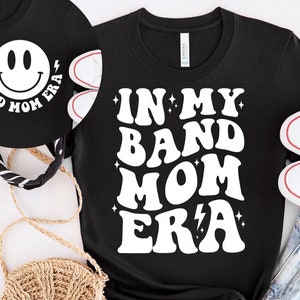 Pu&ograve; includere: Maglietta nera con testo bianco che recita "In My Band Mom Era" in un font retr&ograve;. Il testo &egrave; circondato da piccole stelle bianche e un fulmine.