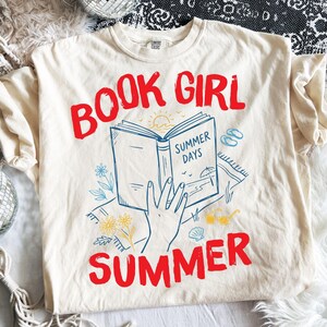 Op de afbeelding: Een crèmekleurig t-shirt met een grafische print met de rode tekst BOOK GIRL SUMMER. Het ontwerp toont een hand die een open boek vasthoudt met het label SUMMER DAYS, omringd door blauwe lijntekeningen van bloemen, een strand en een zonnebril.