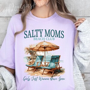 May include: A lavender t-shirt with the text "Salty Moms Beach Club" and "Girls Just Wanna Have Sun" printed on it. The shirt features a graphic of two beach chairs under a beach umbrella.