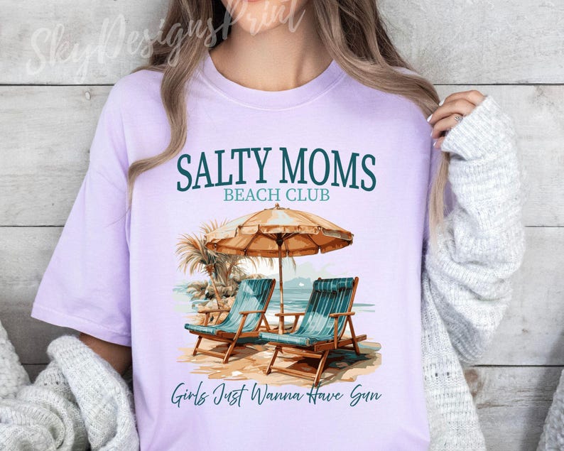 May include: A lavender t-shirt with the text "Salty Moms Beach Club" and "Girls Just Wanna Have Sun" printed on it. The shirt features a graphic of two beach chairs under a beach umbrella.