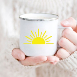 May include: White enamel mug with a yellow sun graphic. The mug is being held by a person wearing a white sweater.
