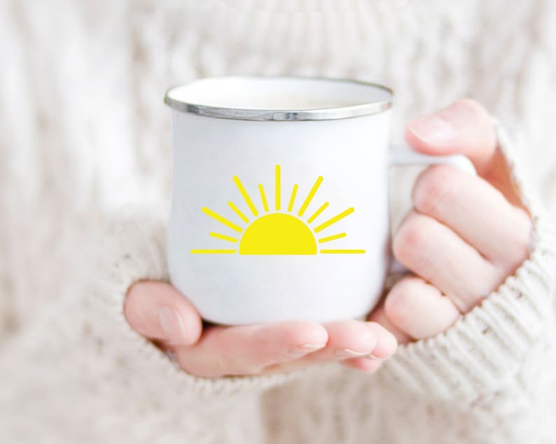 May include: White enamel mug with a yellow sun graphic. The mug is being held by a person wearing a white sweater.