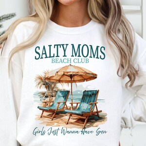 May include: White sweatshirt with a graphic design of two beach chairs under an umbrella with the text "Salty Moms Beach Club" and "Girls Just Wanna Have Sun".
