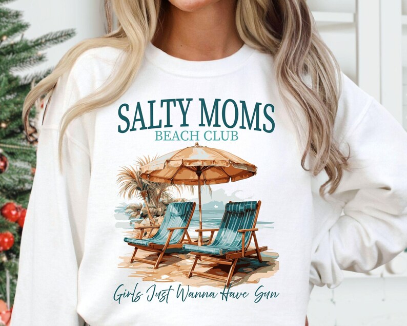 May include: White sweatshirt with a graphic design of two beach chairs under an umbrella with the text "Salty Moms Beach Club" and "Girls Just Wanna Have Sun".
