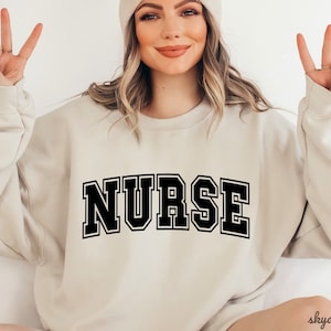 May include: A beige sweatshirt with the word "NURSE" printed in black block letters.