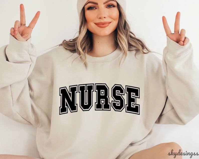 May include: A beige sweatshirt with the word "NURSE" printed in black block letters.