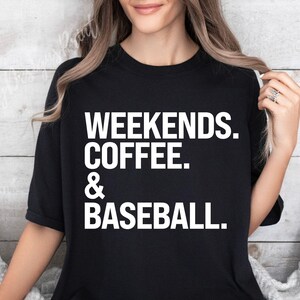 May include: Black t-shirt with the words "WEEKENDS. COFFEE. & BASEBALL." in large white block letters. The shirt is a crew neck and appears to be made of a soft material. The shirt is being worn by a person.