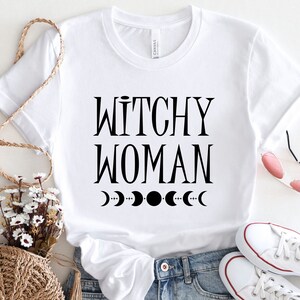 May include: White t-shirt with black text that reads "Witchy Woman" and a row of black crescent moons below the text.
