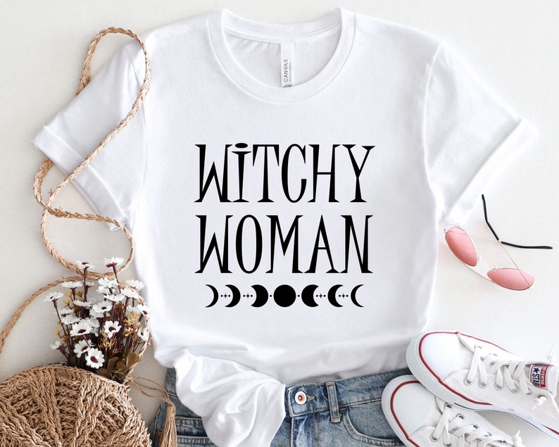May include: White t-shirt with black text that reads "Witchy Woman" and a row of black crescent moons below the text.