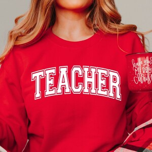 May include: A red sweatshirt with the word "TEACHER" in large, white, collegiate-style lettering. A red mug with white text that reads "Have yourself a Merry Little Christmas" is held in the hand.