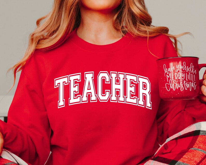 May include: A red sweatshirt with the word "TEACHER" in large, white, collegiate-style lettering. A red mug with white text that reads "Have yourself a Merry Little Christmas" is held in the hand.