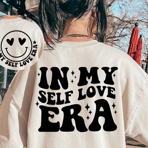 May include: White sweatshirt with black text that reads "In My Self Love Era" in a retro font. The text is surrounded by small stars.