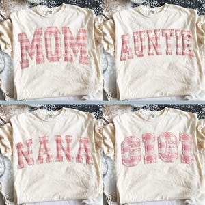 May include: Four cream-coloured t-shirts, each with a different name in pink gingham letters: MOM, AUNTIE, NANA, and GIGI. The letters are stitched onto the shirts. The shirts are displayed on a patterned surface.