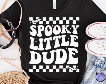 May include: Black t-shirt with the text "SPOOKY LITTLE DUDE" in white, retro-style lettering. The design includes small star accents and bat silhouettes. A pair of white sneakers and denim shorts are visible.