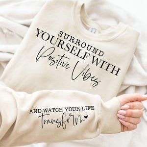 May include: Beige sweatshirt with the text "SURROUND YOURSELF WITH Positive Vibes" on the front. The sleeve displays the text "AND WATCH YOUR LIFE Transform" with a heart symbol. The sweatshirt is folded on a white surface.