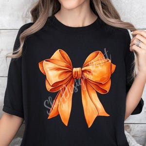 Pode incluir: Uma camiseta preta com um grande desenho de la&ccedil;o laranja. O la&ccedil;o &eacute; feito de cetim e tem uma forma cl&aacute;ssica.