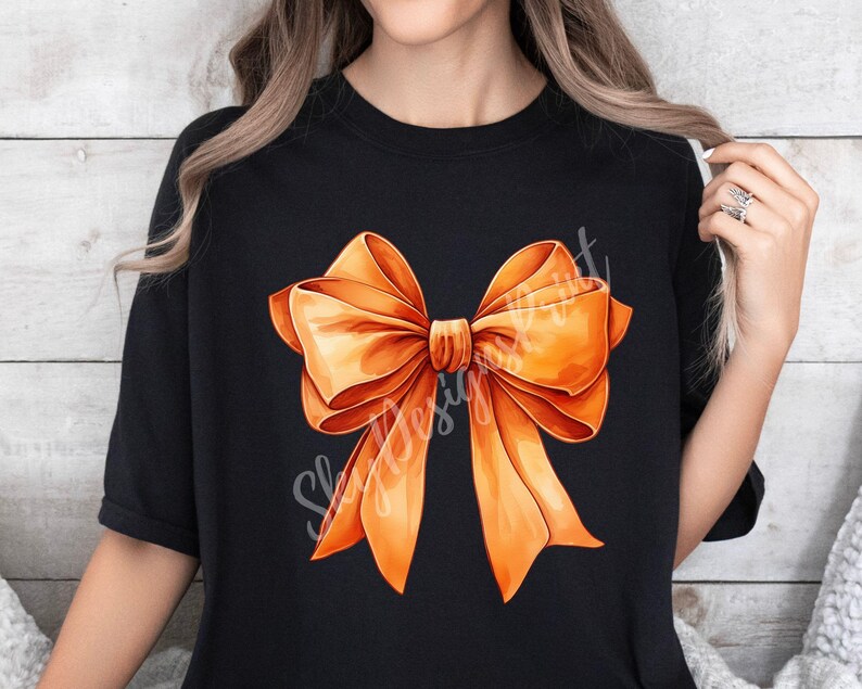 Pode incluir: Uma camiseta preta com um grande desenho de la&ccedil;o laranja. O la&ccedil;o &eacute; feito de cetim e tem uma forma cl&aacute;ssica.