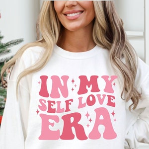 May include: White sweatshirt with pink lettering that reads "In My Self Love Era".
