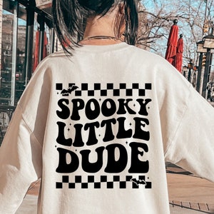 Peut inclure: Sweat-shirt cr&egrave;me avec l'inscription noire "SPOOKY LITTLE DUDE", agr&eacute;ment&eacute;e de chauves-souris et d'&eacute;toiles. Le motif comprend un motif &agrave; carreaux noir et blanc en haut et en bas. V&ecirc;tement sur le th&egrave;me d'Halloween.