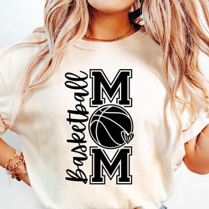 Może przedstawiać: Biała koszulka z czarnym napisem "Basketball Mom" z grafiką koszyk&oacute;wki i serca.