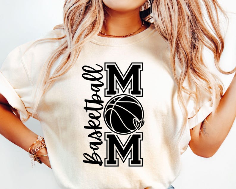 Może przedstawiać: Biała koszulka z czarnym napisem "Basketball Mom" z grafiką koszyk&oacute;wki i serca.