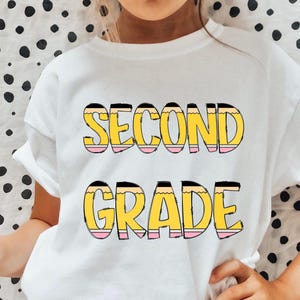 May include: White t-shirt with the words "Second Grade" in yellow and pink letters that look like pencils.