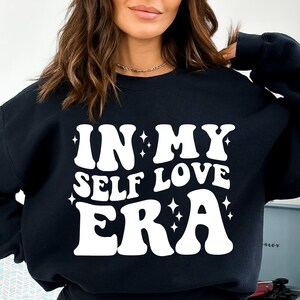 May include: Black sweatshirt with white text that reads "IN MY SELF LOVE ERA" with small white stars around the text.