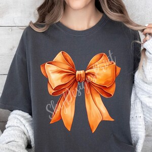 Pode incluir: Uma camiseta cinza escura com um grande design de la&ccedil;o laranja. O la&ccedil;o &eacute; feito de fita de cetim e tem uma forma cl&aacute;ssica.