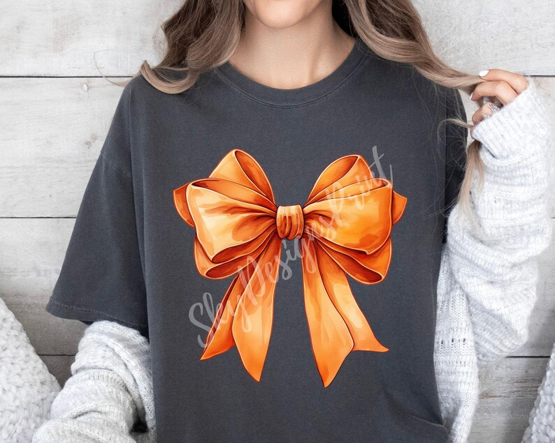 Pode incluir: Uma camiseta cinza escura com um grande design de la&ccedil;o laranja. O la&ccedil;o &eacute; feito de fita de cetim e tem uma forma cl&aacute;ssica.