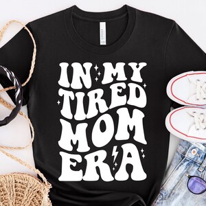 以下が含まれることがあります： 白い文字で「In My Tired Mom Era」と書いてある黒いTシャツ。星と稲妻のグラフィックが描かれている。