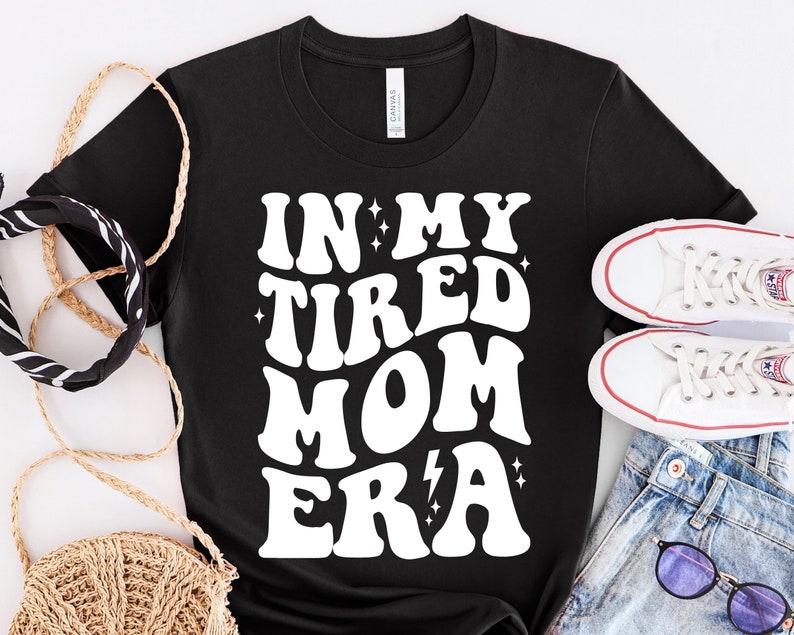以下が含まれることがあります： 白い文字で「In My Tired Mom Era」と書いてある黒いTシャツ。星と稲妻のグラフィックが描かれている。