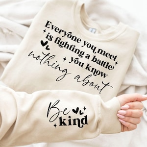 以下が含まれることがあります： 「Everyone you meet is fighting a battle you know nothing about」と「Be kind」の黒い文字がプリントされたベージュのスウェットシャツ。クルーネックと長袖のデザインです。