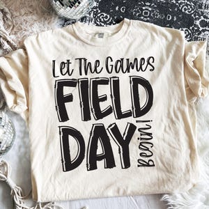 May include: Cream-colored t-shirt with the text "Let The Games FIELD DAY Begin!" in black. The shirt has a crew neck and short sleeves. The text is in a bold, playful font.